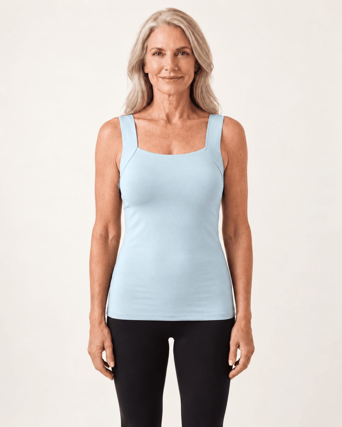 A woman modeling a light blue scoop-neck tank top with wide shoulder straps and black leggings, standing against a plain off-white studio background.