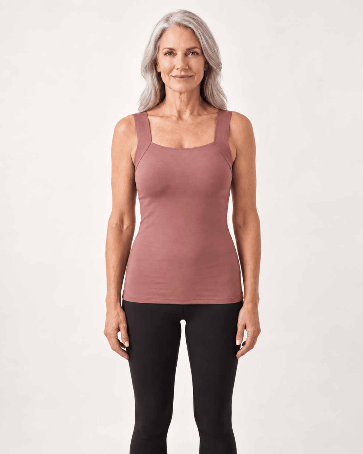 A woman with gray hair wearing a dusty rose ribbed tank top and black leggings, standing against a light gray studio background.
