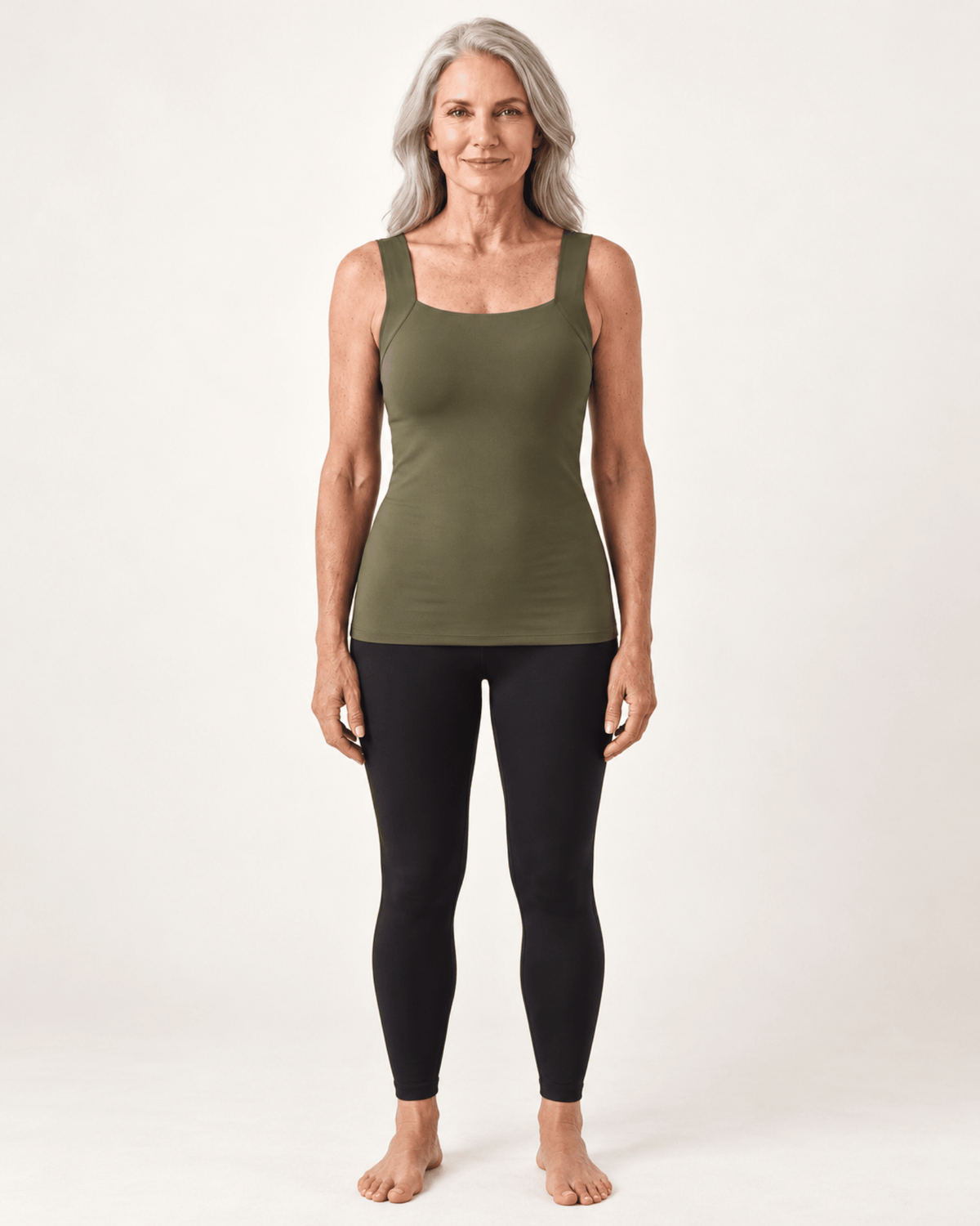 A woman with gray hair stands barefoot against a plain light background, wearing an olive green sleeveless tank top and black leggings.