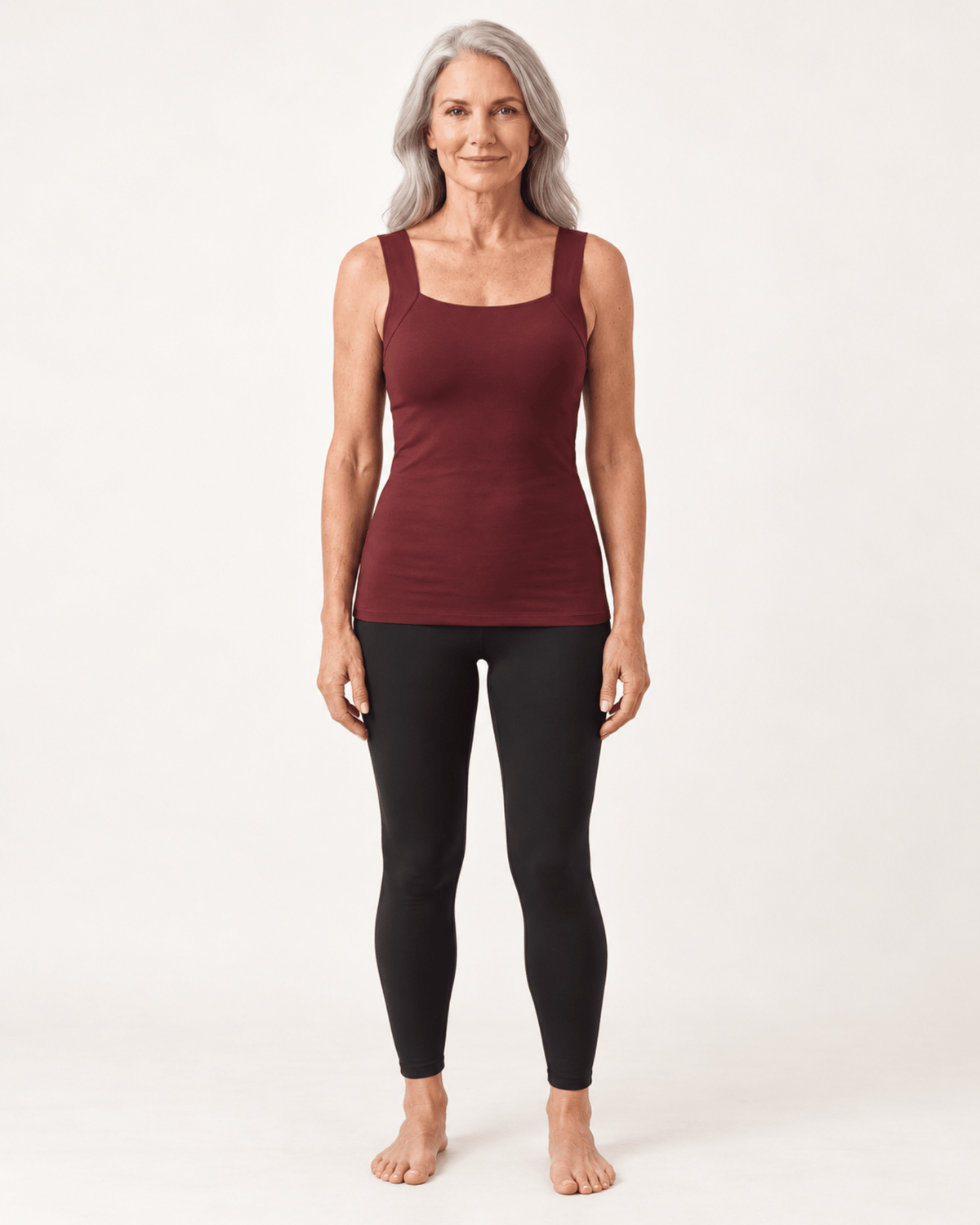 A woman with gray hair stands barefoot against a plain light background, wearing a sleeveless scoop-neck tank top in burgundy and black leggings.