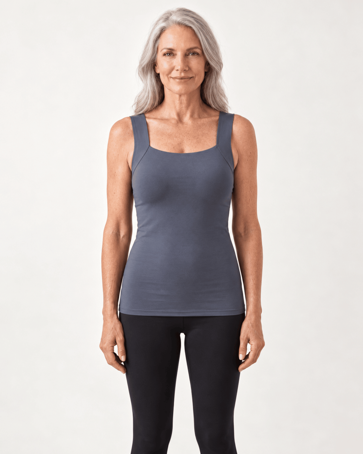 A woman with gray hair wearing a slate blue-gray ribbed tank top and black leggings, standing against a light gray studio background.