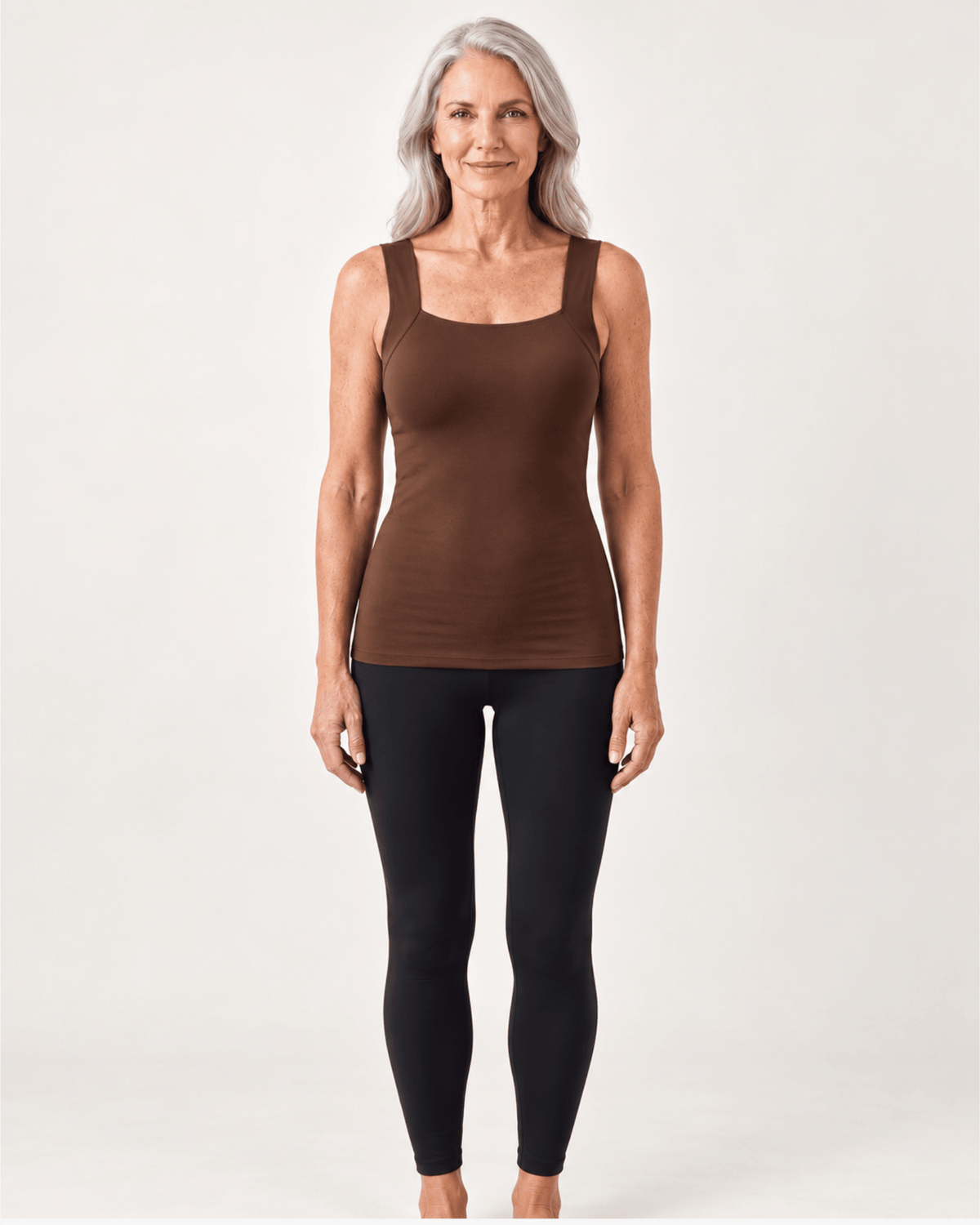 An older woman stands against a light gray studio background wearing a fitted brown tank top and black leggings.