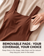 A brown camisole-style top laid flat on a beige textured blanket, with a hand placing a light beige removable bra pad into one of the shoulder cutouts. Large text at the bottom reads: 'REMOVABLE PADS - YOUR COVERAGE, YOUR CHOICE' with smaller supporting text below.