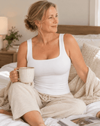 A woman sitting on a bed in a bedroom, wearing a white tank top and beige lounge pants while holding a ceramic mug.