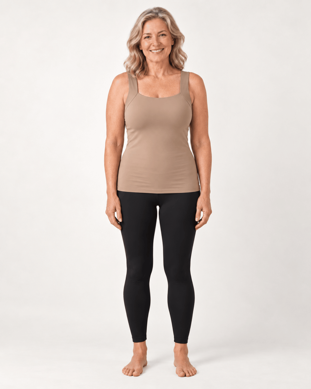 A woman stands barefoot against a light gray studio background, wearing a fitted scoop-neck tank top in taupe and black leggings.