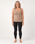 A woman stands barefoot against a light gray studio background, wearing a fitted scoop-neck tank top in taupe and black leggings.