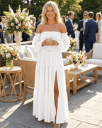 Woman in a white dress standing outdoors at a social event with floral arrangements and people in the background.